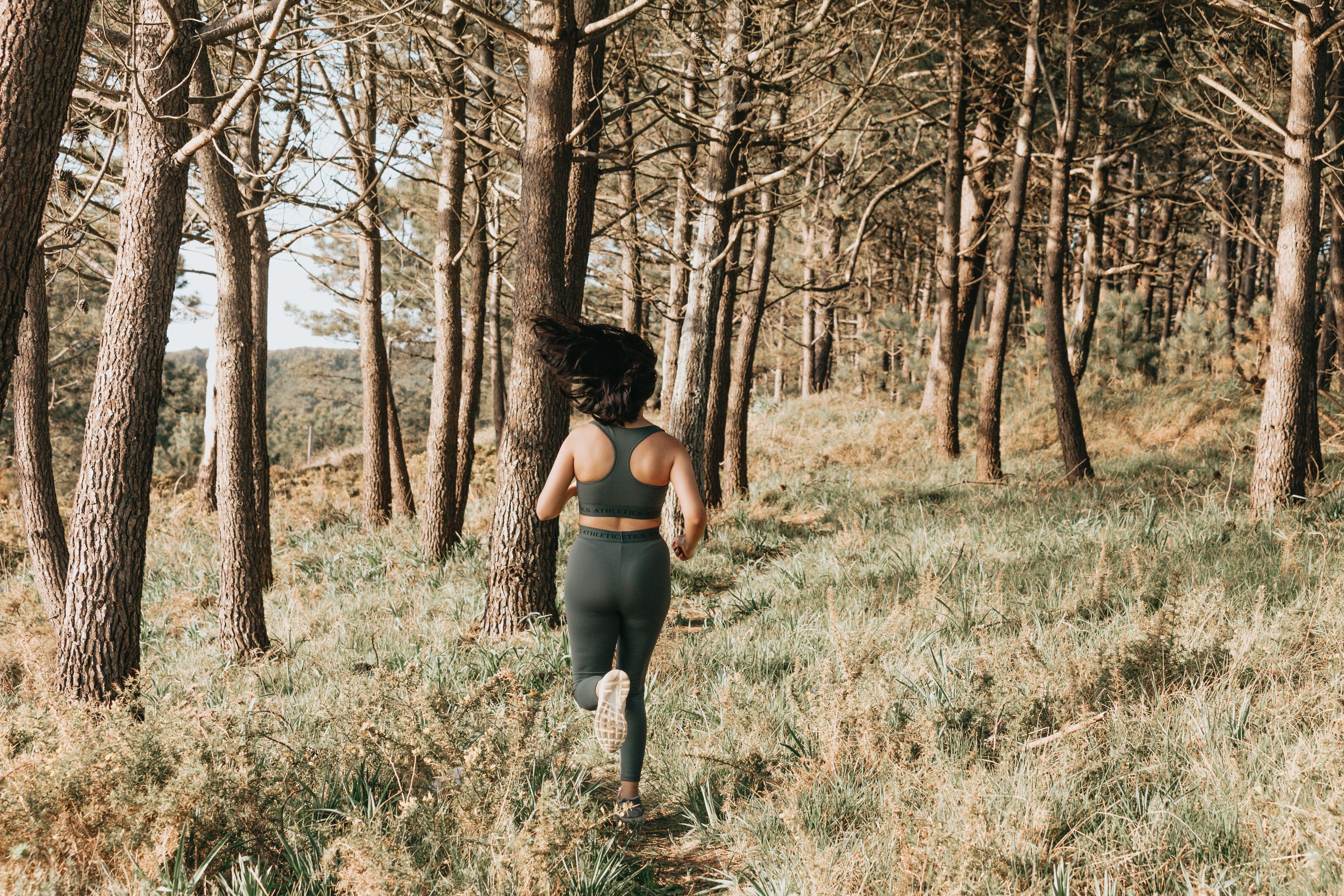 This is an image of a forest on a sunny day. A woman in green matching sports wear is running away from the camera down a trail.